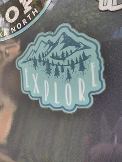 A teal and dark teal sticker with a mountain range and forest design. The word 'EXPLORE' is written in a stylized font below the image.
