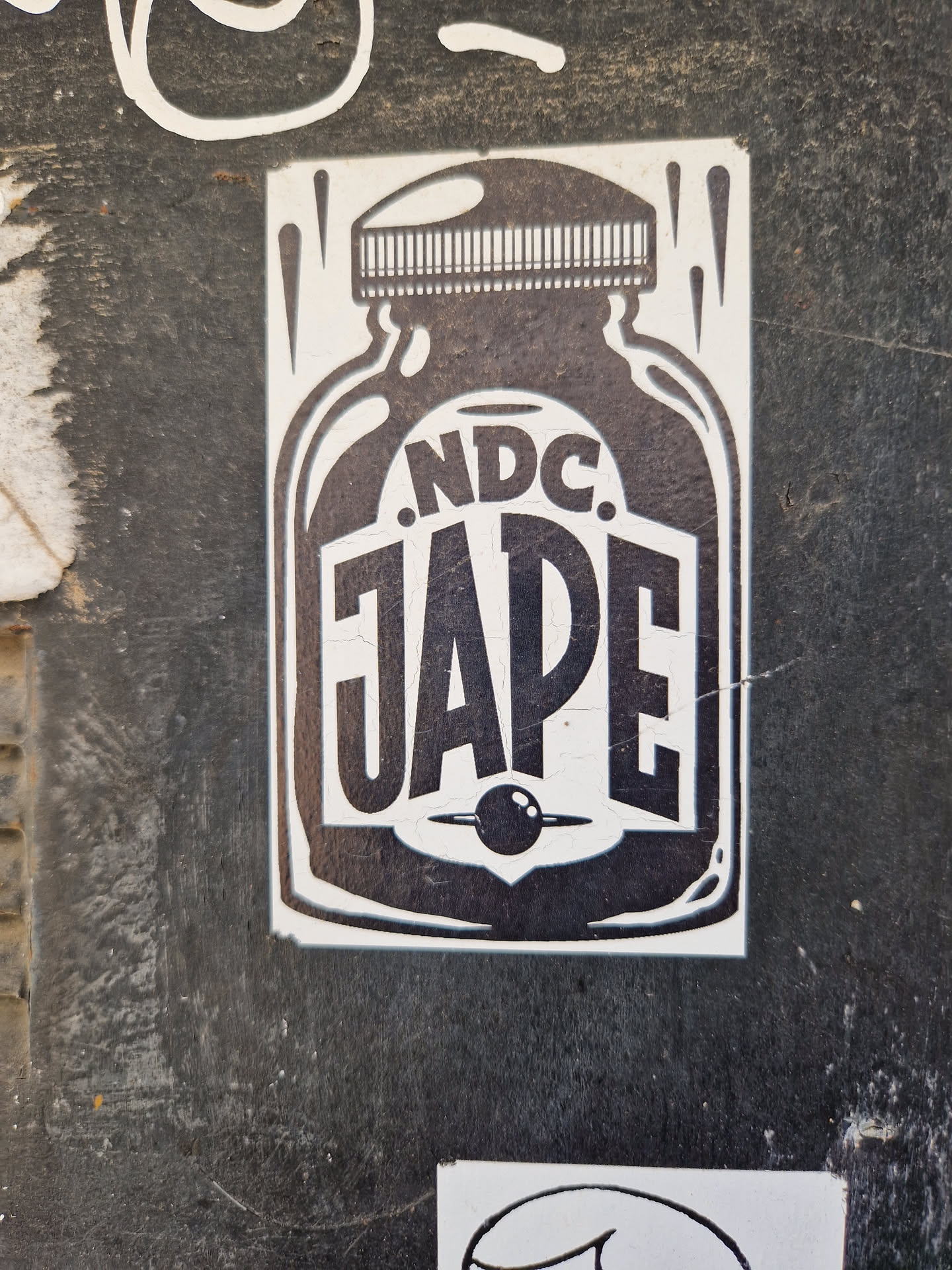 A black and white sticker depicting a jar with the text ".NDC. JAPE" on it. The design is simple yet striking, with bold lettering and a small circular element at the bottom of the jar.