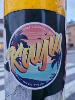 Round sticker with a beach scene and the word "Kayu" in a stylized font.  The text "LOS MA" is repeated around the edge of the sticker, along with the website URL.