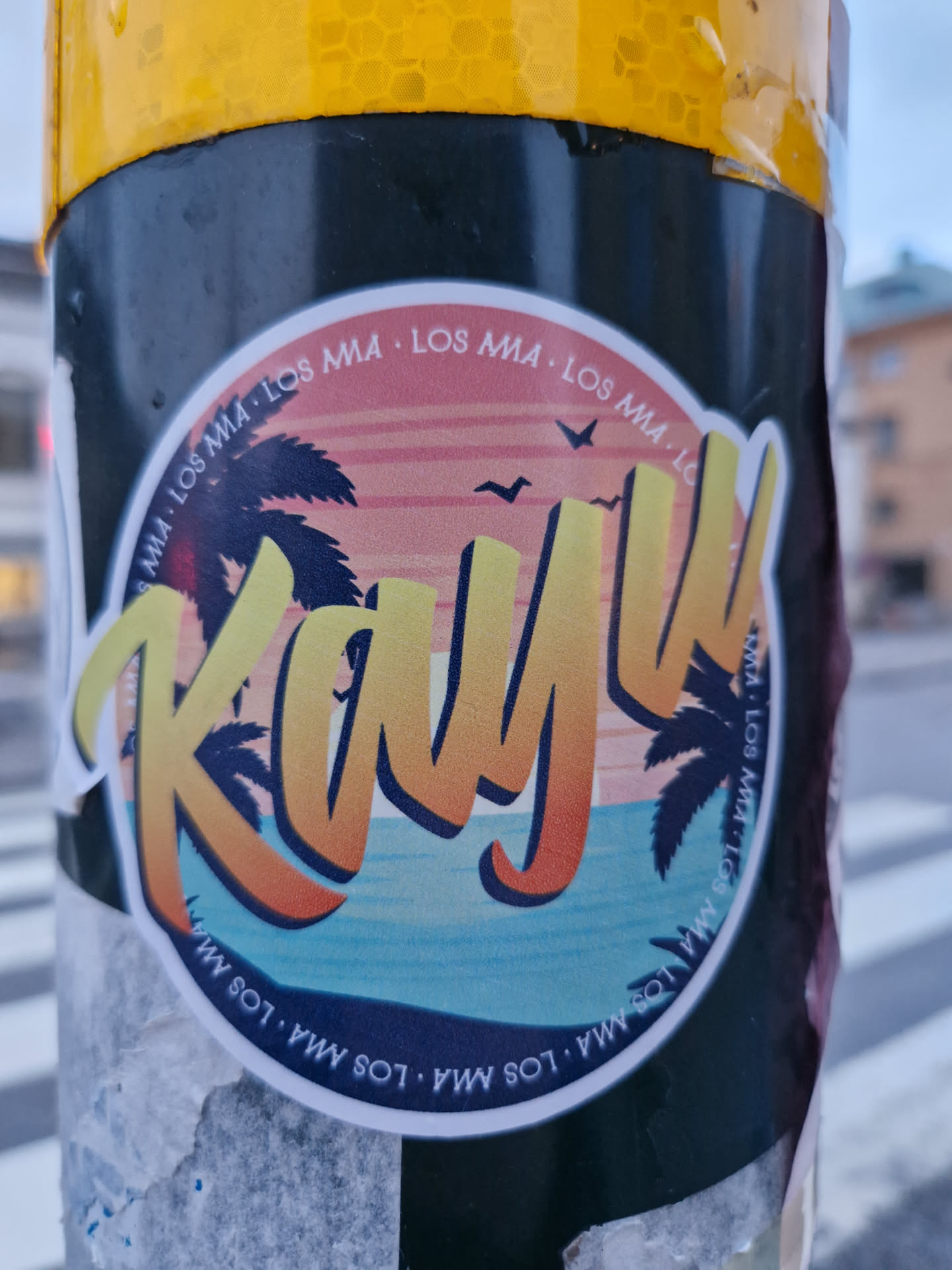 Round sticker with a beach scene and the word "Kayu" in a stylized font.  The text "LOS MA" is repeated around the edge of the sticker, along with the website URL.