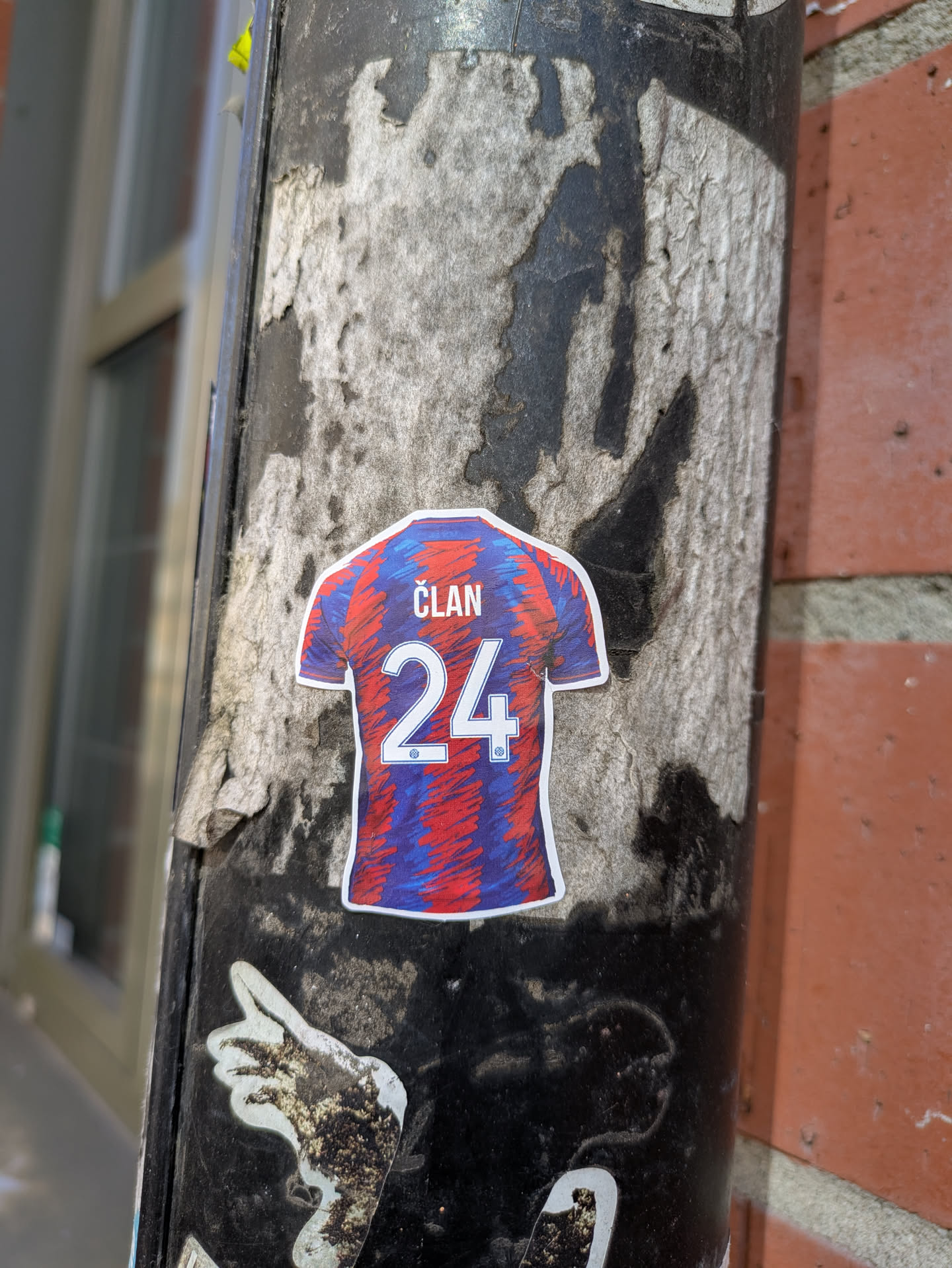 This is a sticker featuring a soccer jersey with a modern design, showcasing a blend of red and blue hues. The name "ČLAN" is printed across the upper back, just above the number "24". The sticker is placed on a weathered black pole.