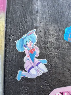 A sticker features an anime-style girl with blue hair and bunny ears, wearing a blue bodysuit with a red bow. The sticker is placed on a textured gray surface.