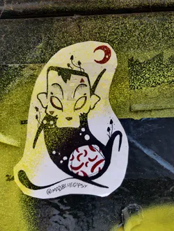 A whimsical sticker featuring an alien-like creature with large eyes, adorned with small plants and a crescent moon. The character has a dark silhouette with red and white patterned accents. The artist's Instagram handle is visible in the lower right corner.