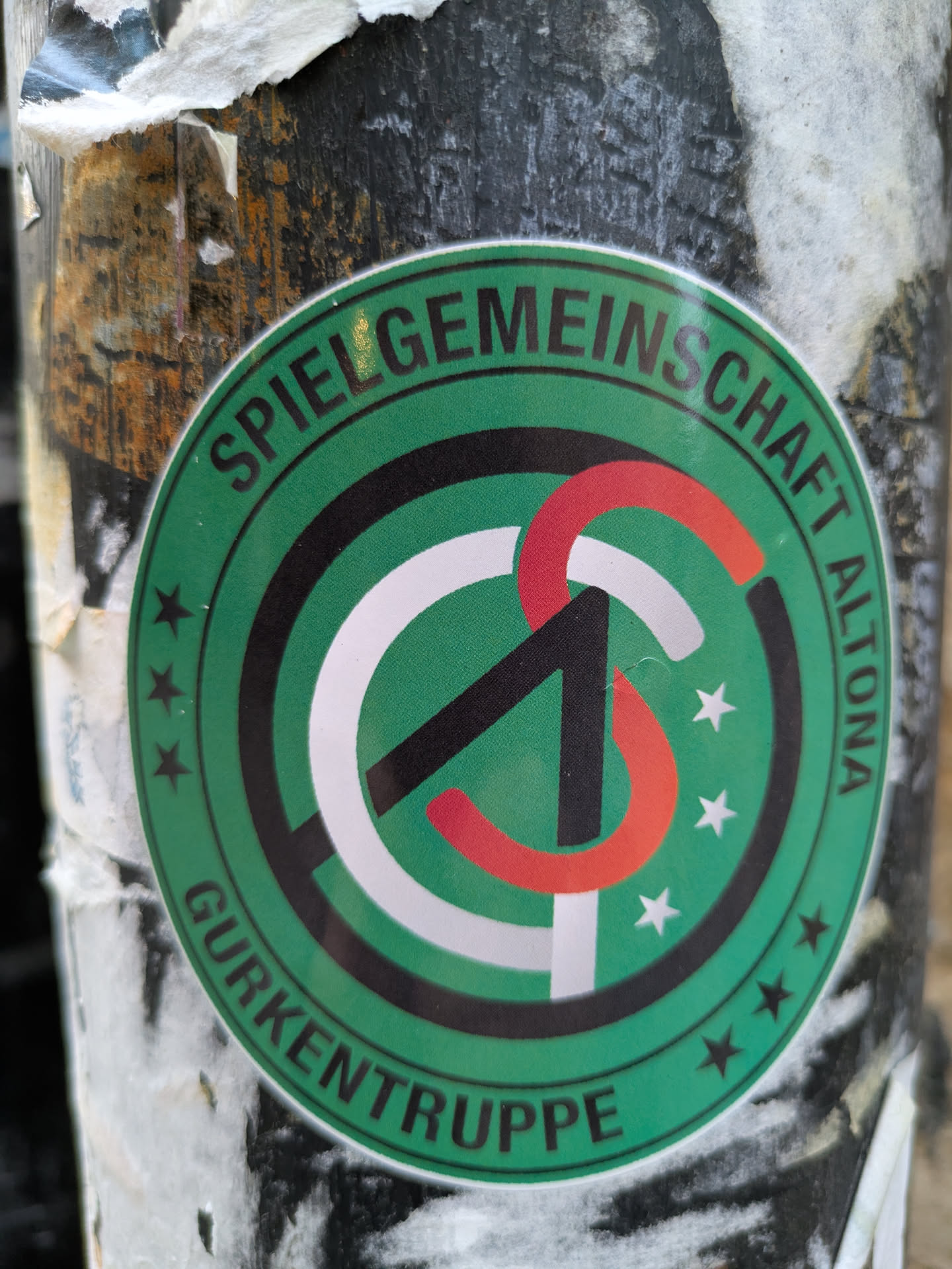 Round green sticker with a logo and text. The logo is composed of a stylized letter 'S' in black, white and red. The text around the logo reads 'Spielgemeinschaft Altona Gurkentruppe'.