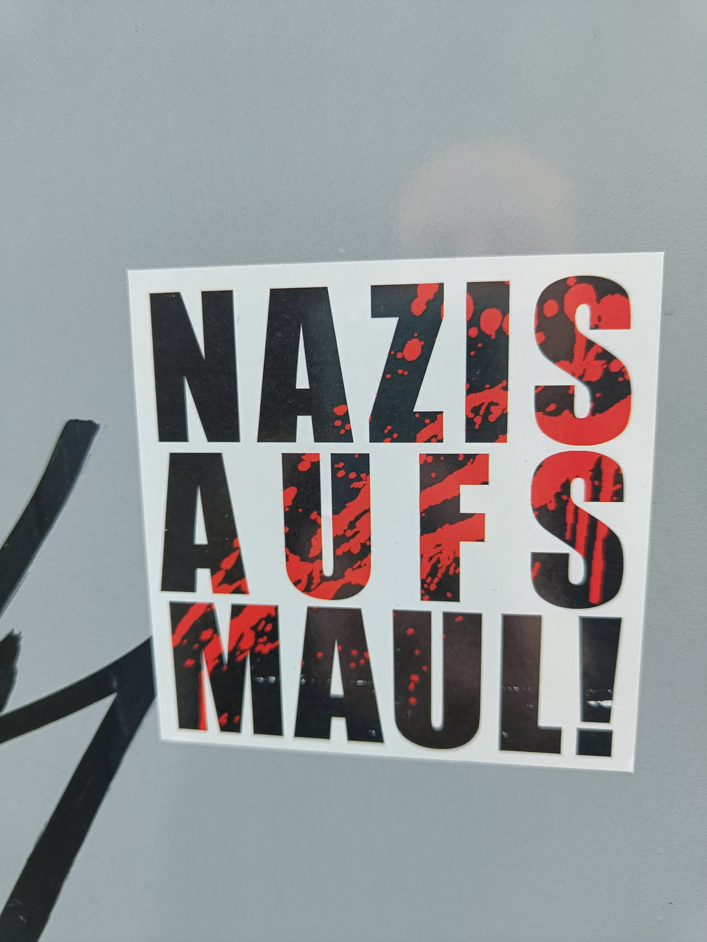A sticker with the text "Nazis Auf's Maul!" in black and red. The text is stylized with red splatters behind it. The sticker is square and printed on white paper.
