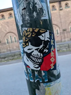 A rectangular sticker featuring a black and white skull profile wearing a gold laurel wreath. The background has diagonal red and blue stripes, with the vertical text 'IKAROS' on the right.