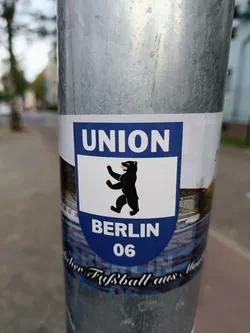 A sticker featuring the logo of Union Berlin, a German football club. The logo shows a bear standing on its hind legs, with the words "UNION" and "BERLIN 06" written above and below it respectively. The sticker also includes some blurry text at the bottom.