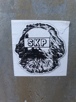 A sticker featuring a black and white illustration of a head, possibly a classical bust, with the letters "SKP" boldly printed across the forehead in a rectangular banner.  The style is reminiscent of woodcut prints. The sticker is affixed to a gray concrete surface.