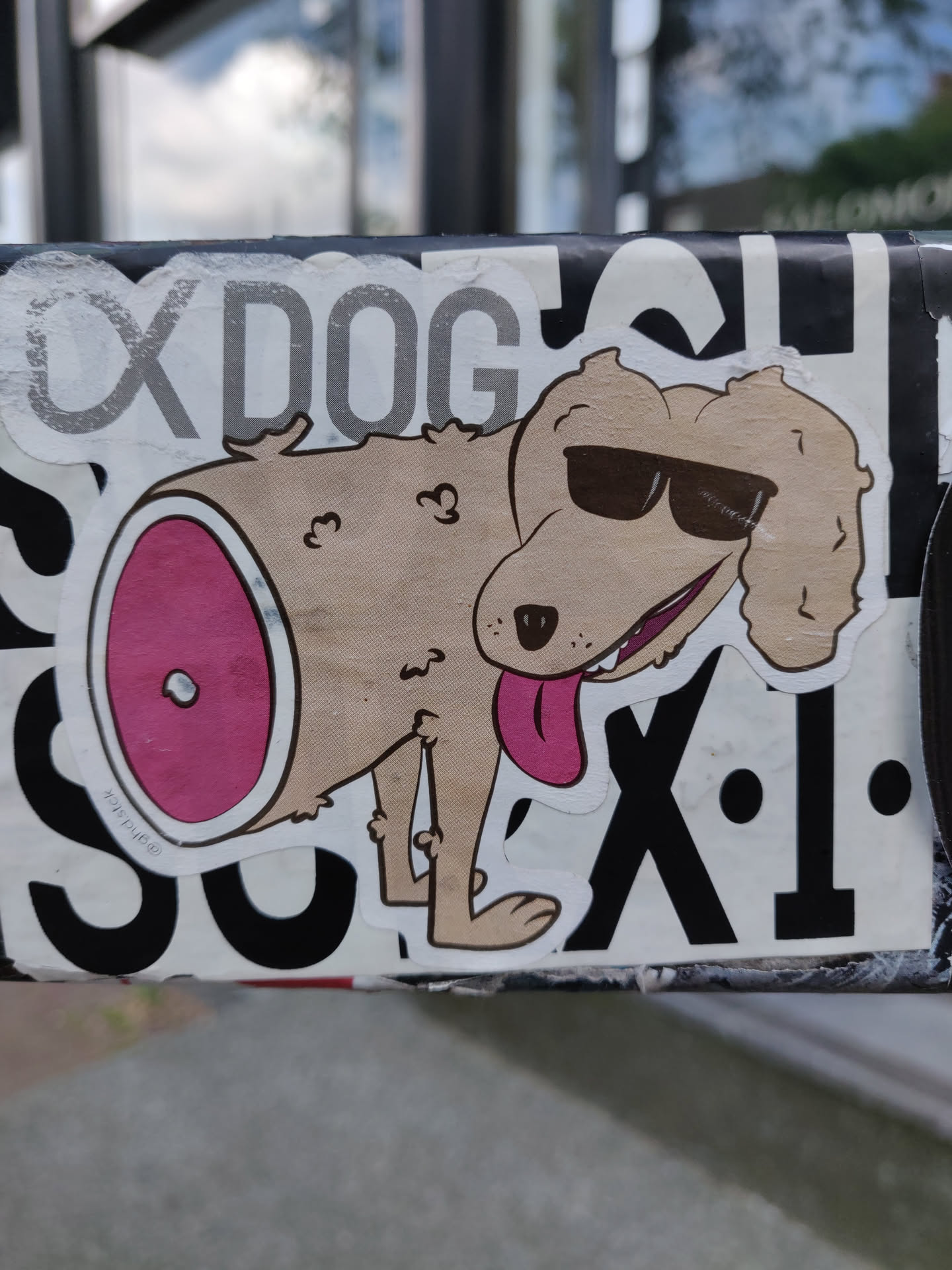 A sticker featuring a cartoon dog wearing sunglasses. The dog has a piece of meat on its behind. The sticker is on a pole outside.
