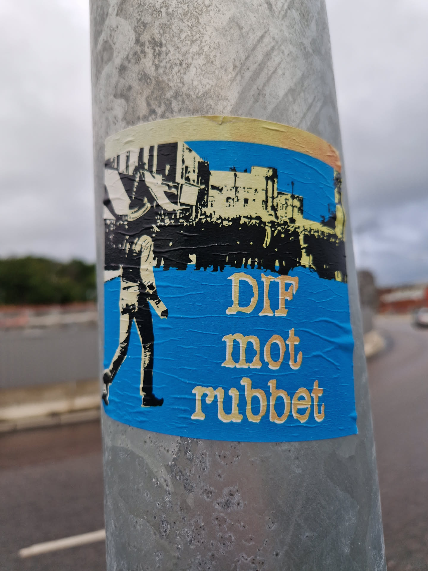 A blue sticker with a black and white illustration of a person walking in front of a crowd and buildings. The text 'DIF mot rubbet' is written in a serif font below the illustration. The sticker is affixed to a metal pole.