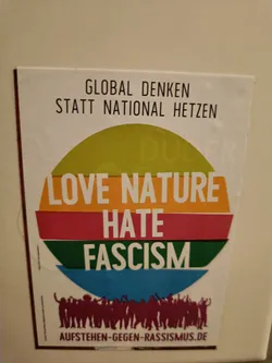 Sticker promoting anti-fascist and environmentalist messages. Features a circular design with a rainbow color scheme and silhouettes of people raising their hands in celebration. The text also includes the German phrase "Global Denken statt national Hetzen," which translates to "Think globally, not nationally hate."  A website address is present at the bottom. 