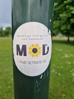 A worn sticker on a green pole. The sticker features a sunflower graphic with the letters MOD surrounding it.  Text on the sticker reads "Människorättigheter och Demokrati" and "PARTIETMOD.SE".