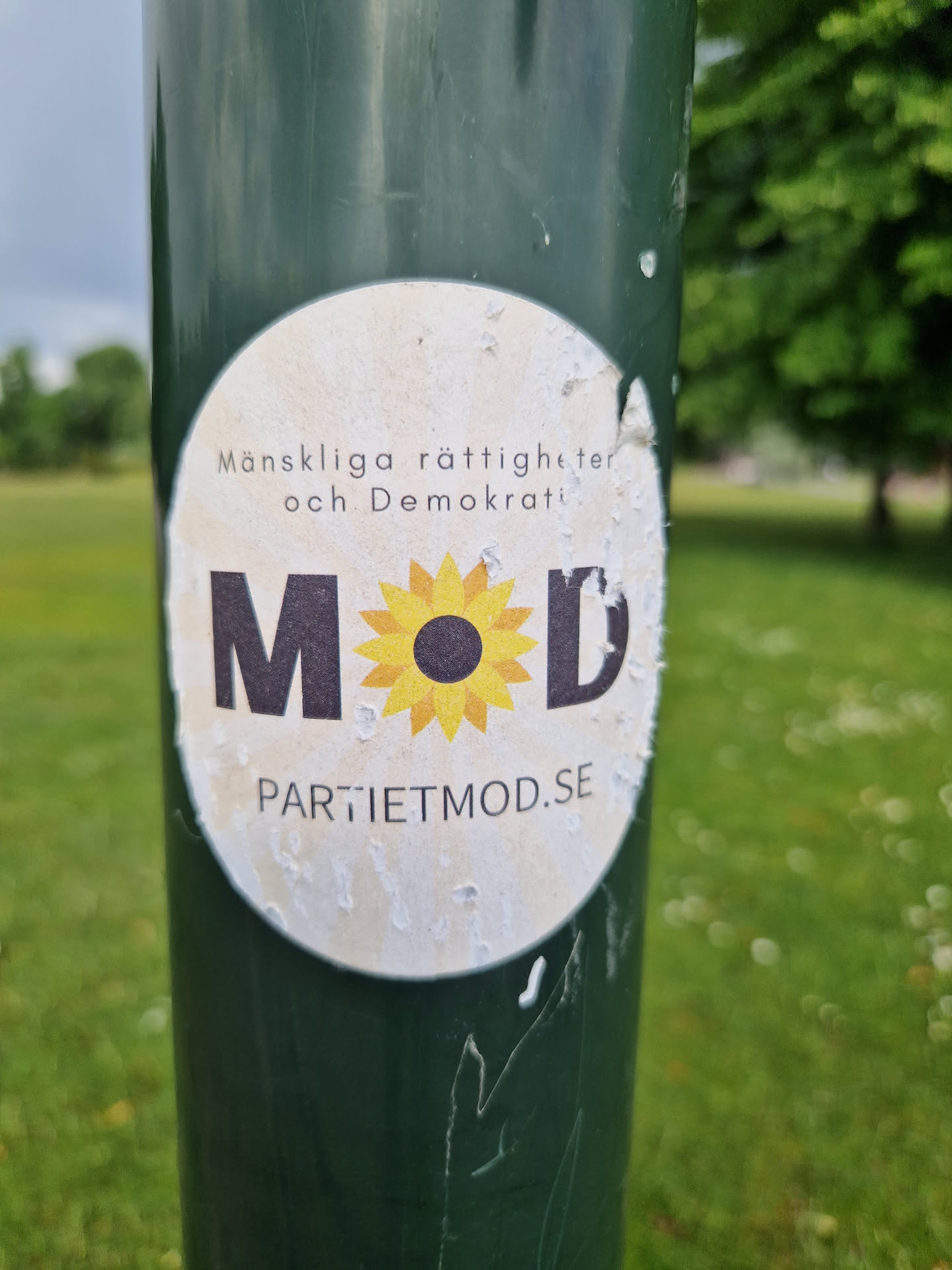 A worn sticker on a green pole. The sticker features a sunflower graphic with the letters MOD surrounding it.  Text on the sticker reads "Människorättigheter och Demokrati" and "PARTIETMOD.SE".