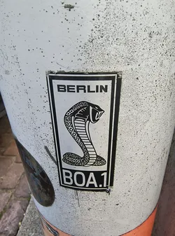 A black and white sticker featuring a cobra with the text 'BERLIN' above and 'BOA.1' below the cobra on a white background with a black border. The cobra has a detailed diamond pattern on its body and is poised to strike. The sticker is affixed to a light-colored, cylindrical surface.