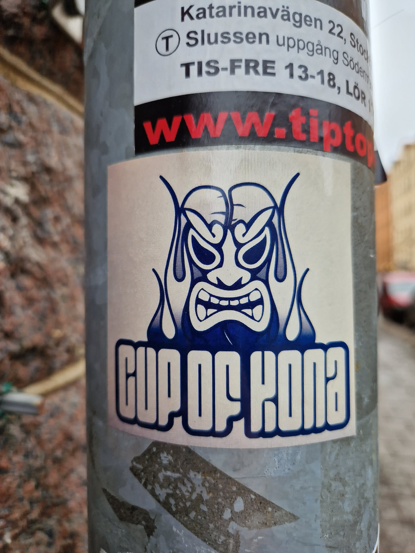 A sticker featuring a stylized, angry-looking face, possibly inspired by a Tiki mask, set within blue flames. Below it, the words "Cup of Kona" are prominently displayed. The sticker appears to be newly applied.