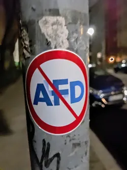 A sticker with a red circle and a diagonal red line crossing out the letters AFD in blue. The sticker is affixed to a gray pole.