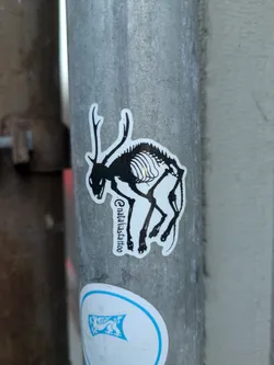 A white die-cut sticker on a gray metal pole features a stylized black image of a deer whose skeletal structure is visible. The sticker has the text "@nataliadstattoo" on its left side. 