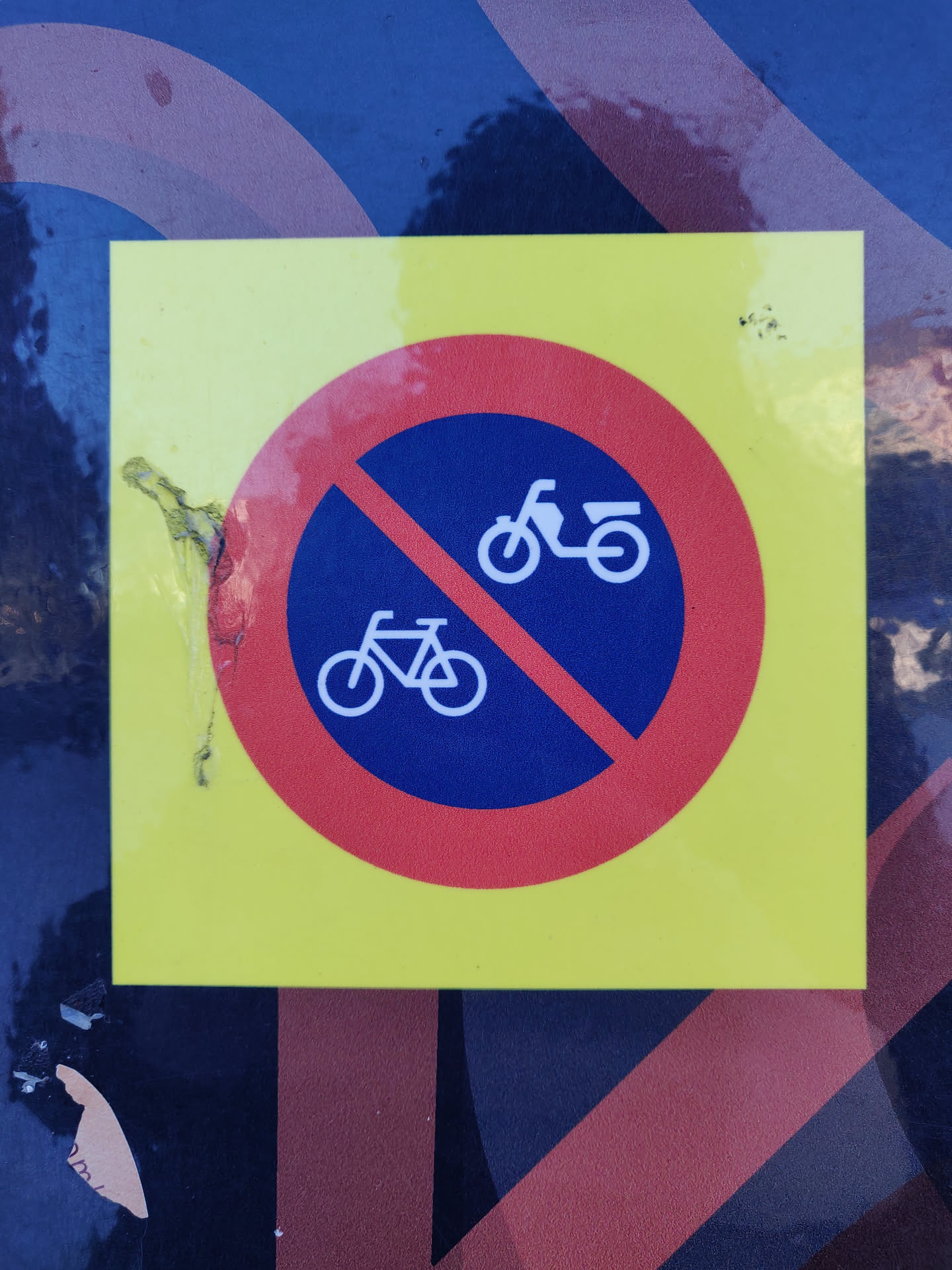 A yellow sticker with a red circle and a diagonal line through the images of a bicycle and a scooter.  The sticker is damaged, showing some peeling at the edges.