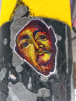 Close-up view of a sticker affixed to a weathered surface. The sticker depicts an expressive face rendered in a colorful, almost painterly style. The colors are bold and the style is slightly abstract, with visible brushstrokes or texture. Water droplets are visible on the sticker's surface. The background is a dark, textured surface with patches of peeling paint.