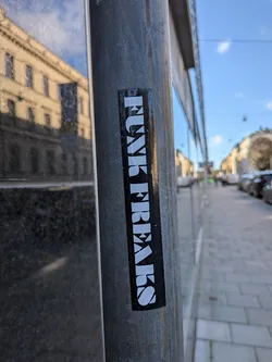 Street sticker A vertical, rectangular sticker with a glossy black background adhered to a gray metal post along a sidewalk. The sticker features the text 'FUNK FREAKS' running vertically from top to bottom in bold, chunky, stylized white uppercase serif lettering.