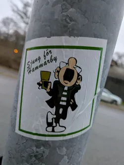 A sticker featuring a cartoon character holding a drink, with text that says "Sjung för Hammarby" (Sing for Hammarby) in Swedish.  The style is reminiscent of vintage cartoons or comic strips. The sticker is affixed to a grey metal pole.