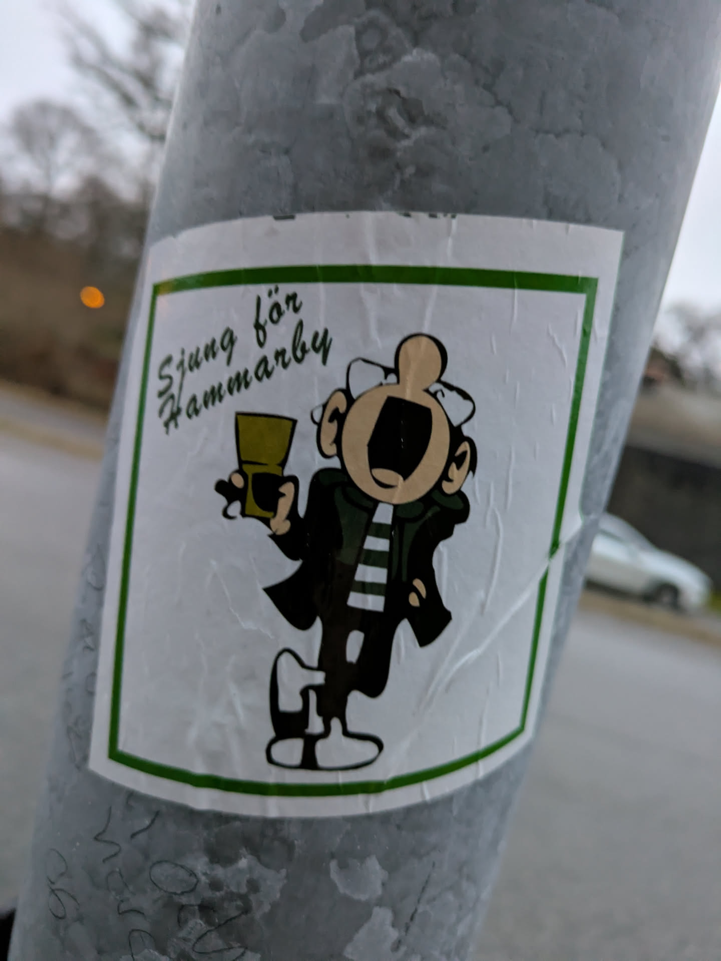 A sticker featuring a cartoon character holding a drink, with text that says "Sjung för Hammarby" (Sing for Hammarby) in Swedish.  The style is reminiscent of vintage cartoons or comic strips. The sticker is affixed to a grey metal pole.