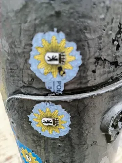 Street sticker Two identical stickers of the German police emblem are affixed to a dark gray pole. The stickers are round, with a light blue background and a yellow sunburst pattern.  In the center is a black and white crest. The pole is textured, and other small stickers are visible in the background.
