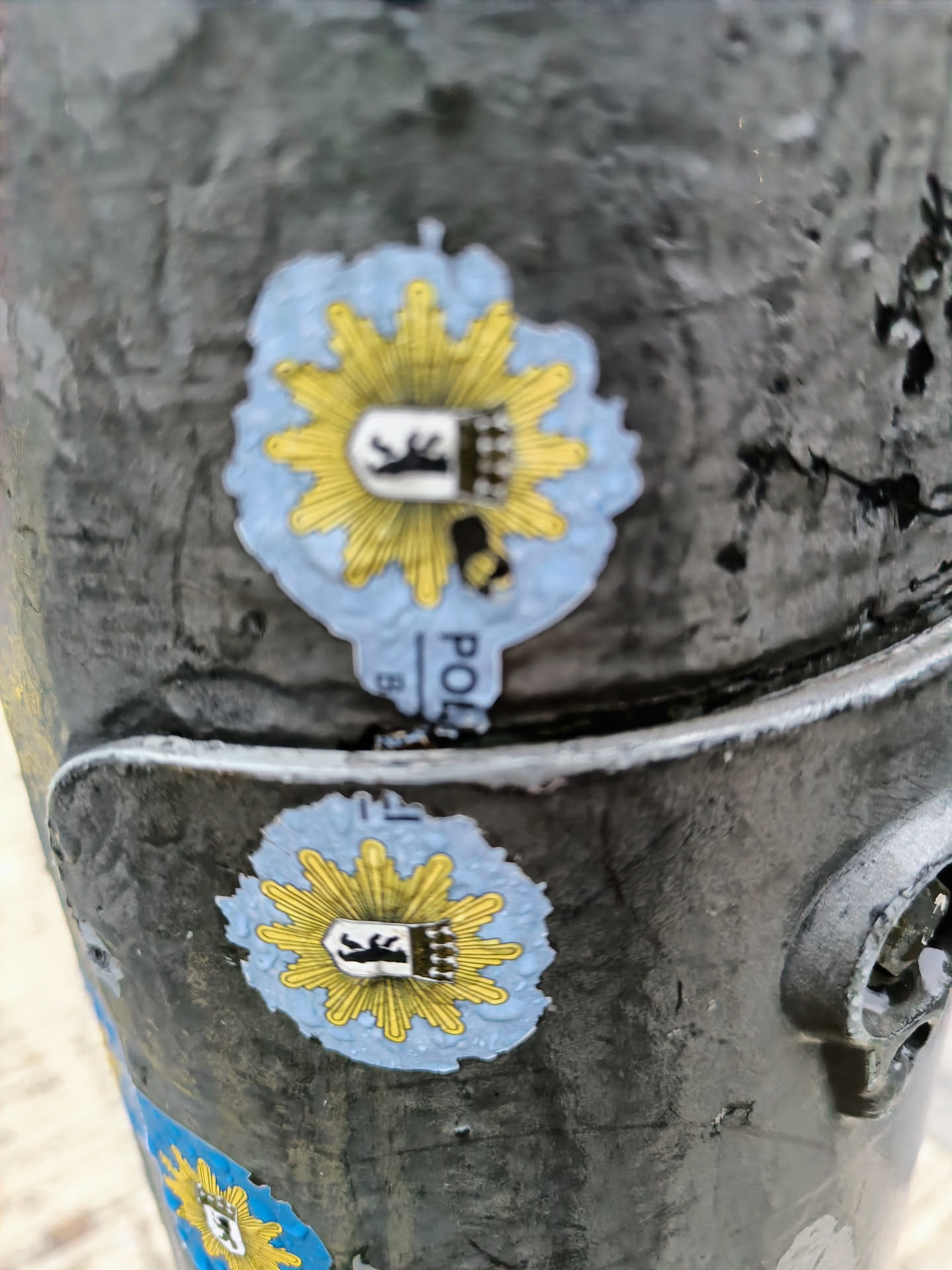 Two identical stickers of the German police emblem are affixed to a dark gray pole. The stickers are round, with a light blue background and a yellow sunburst pattern.  In the center is a black and white crest. The pole is textured, and other small stickers are visible in the background.
