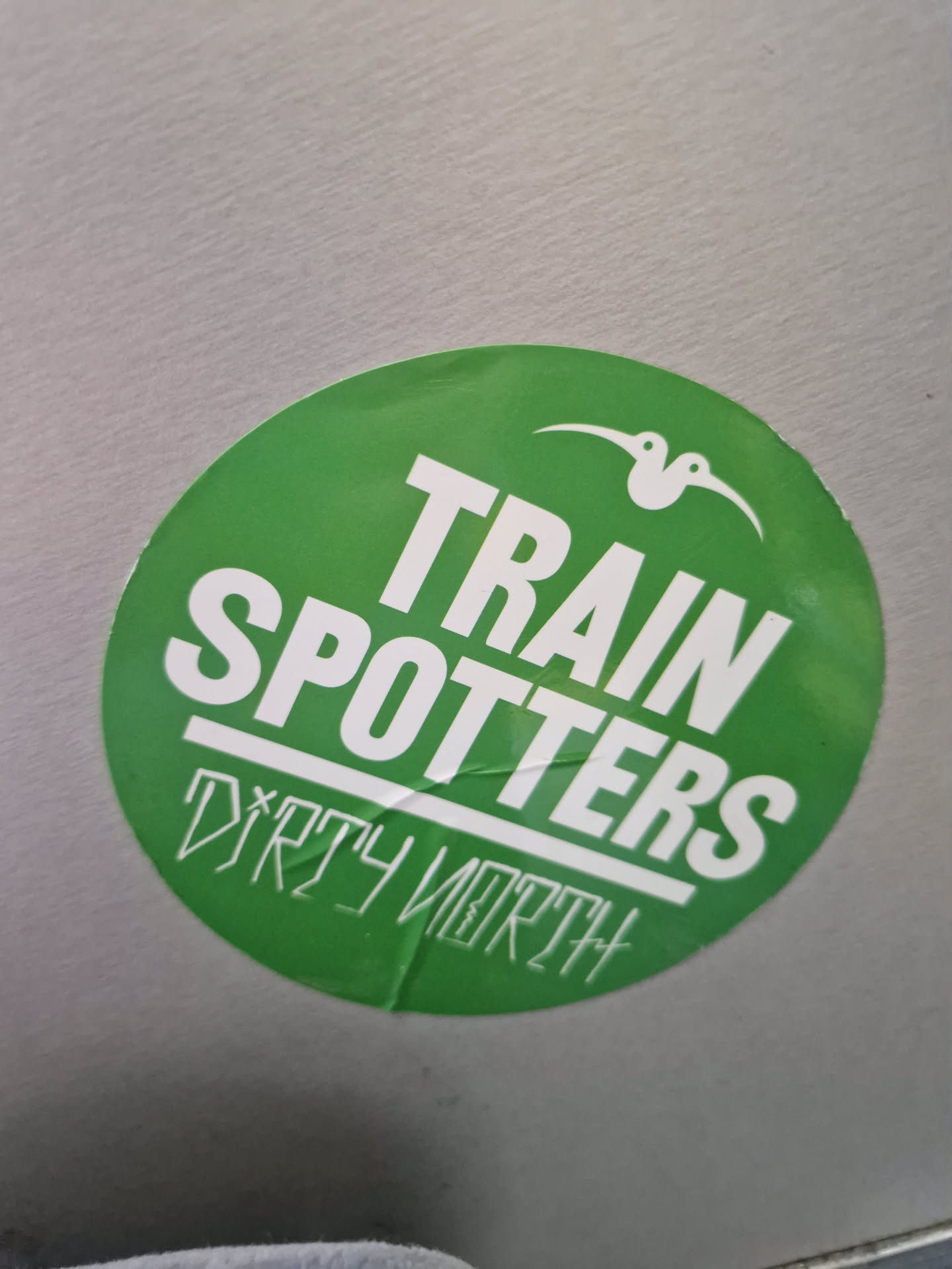 Round green sticker with the text "TRAIN SPOTTERS" in white bold uppercase letters. Underneath, in a smaller, stylized font, it says "DIRTY NORTH". A small bird-like logo is present at the top.