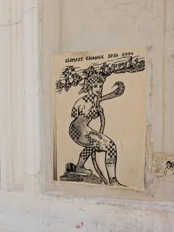 The image shows a black and white poster attached to a wall. The poster presents an illustration of a female figure with a snake draped around her neck. The figure and snake have a checkerboard pattern. Text on the poster reads "CLIMATE CHANGE 2035-2050" and "GRINICY VINTAGE". The overall theme seems to address climate change in a creative and artistic way.