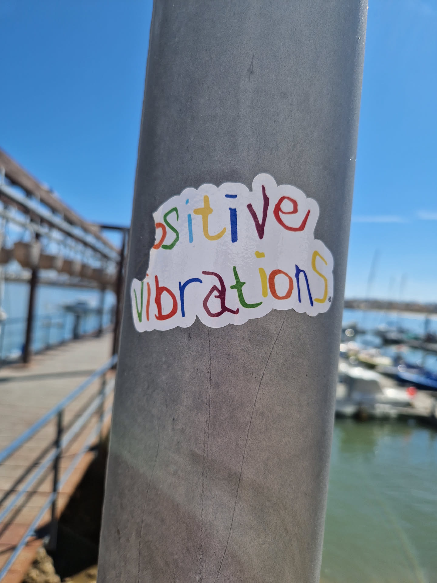 A colorful sticker with the words 'Positive Vibrations' attached to a metal pole near a harbor. The sticker has a hand-drawn, child-like style.