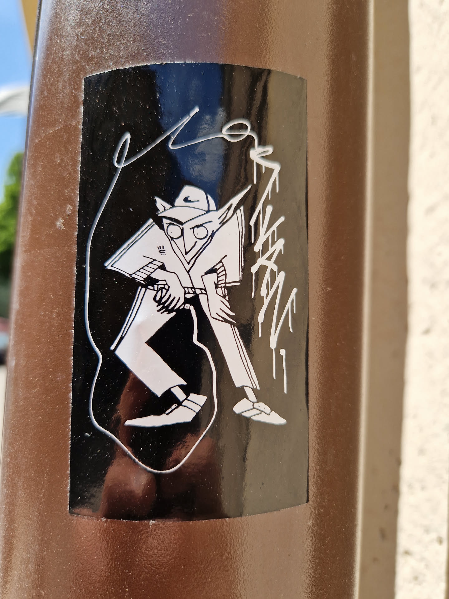 This sticker features a black and white illustration of a character in a hat and coat, possibly inspired by 1920s-era fashion or film noir aesthetics. The style is graphic and slightly cartoonish. The sticker is affixed to a brown pole, likely outdoors.