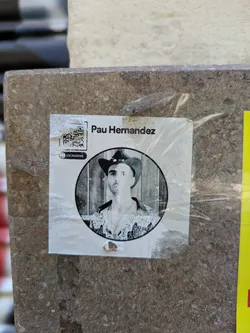 This square sticker has a black and white portrait of Pau Hernandez in a cowboy hat inside a circle. A QR code is in the top left corner with the text "ESCANEAME" below it. The sticker is affixed to a textured, gray surface.
