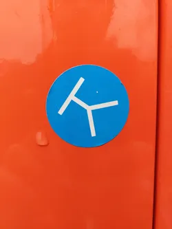 A circular sticker with a blue background and a simple, stylized white design resembling a tripod or a three-legged object.  The sticker is affixed to an orange surface.