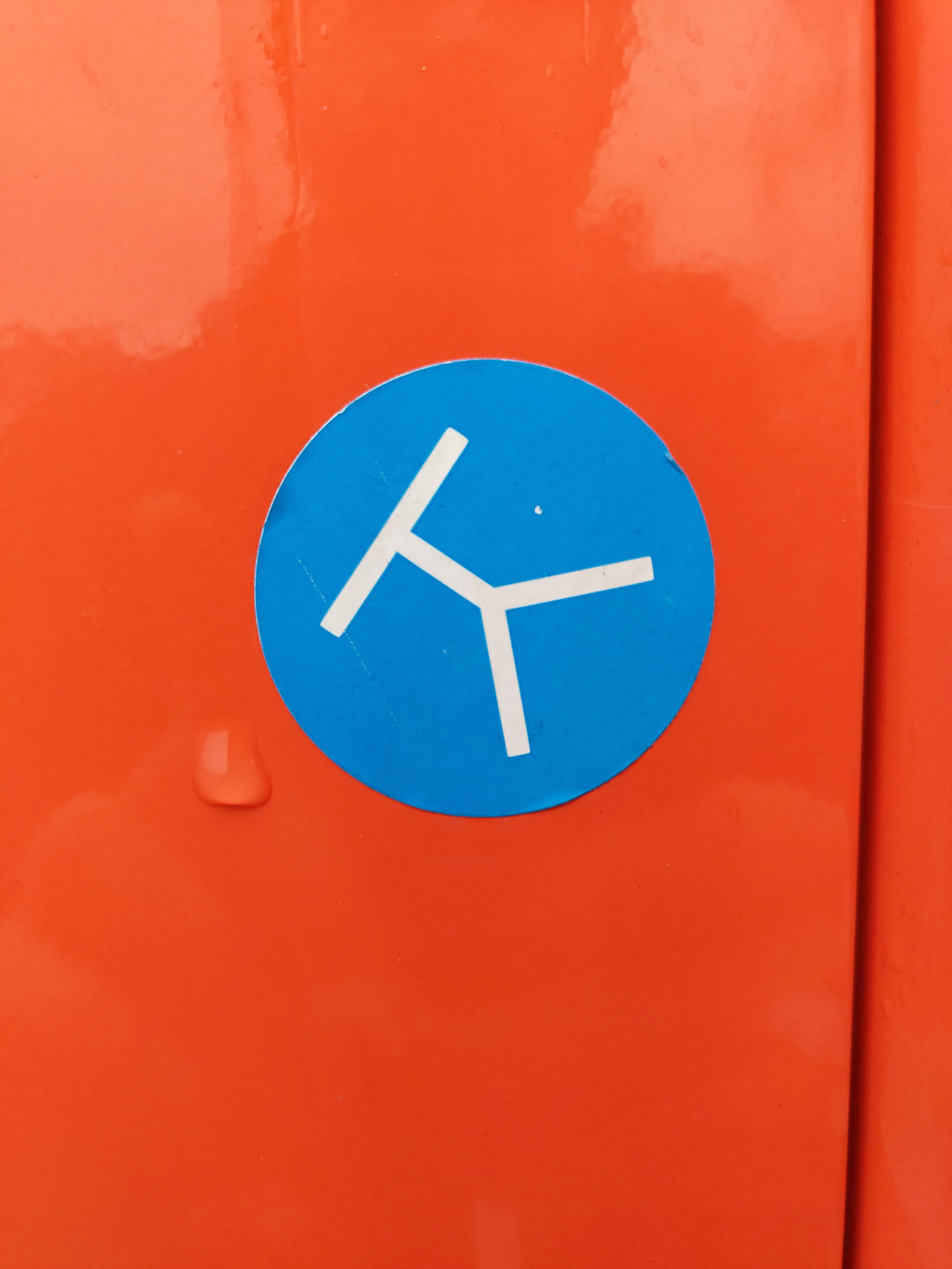 A circular sticker with a blue background and a simple, stylized white design resembling a tripod or a three-legged object.  The sticker is affixed to an orange surface.