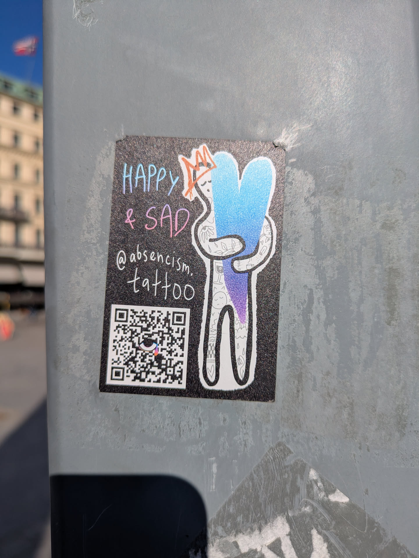 A sticker shows a cartoonish figure hugging a gradient blue and purple heart. The text "Happy & Sad" is written above the figure. Below the figure, the sticker shows the Instagram handle "@absencism.tattoo" and a QR code.