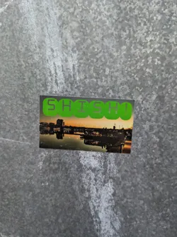 A sticker with the word SHSH written in green letters on top of a picture of a city waterfront at sunset. The city is reflected on the water.