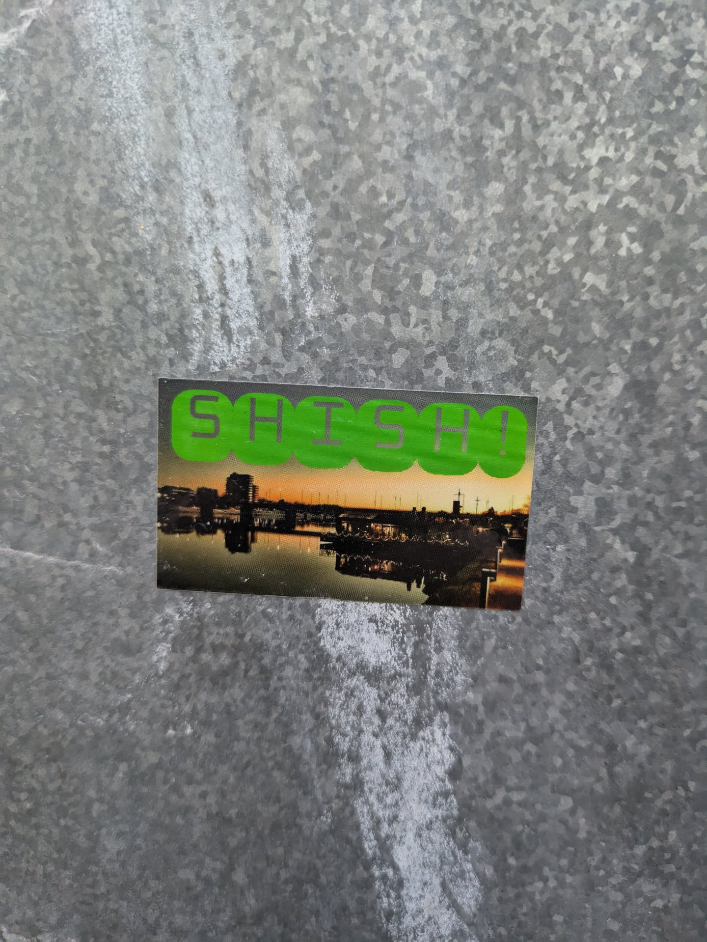 A sticker with the word SHSH written in green letters on top of a picture of a city waterfront at sunset. The city is reflected on the water.