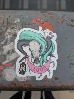 This sticker features a ram skull with turquoise tentacles emerging from it. A pink banner is draped beneath, with the number "1000" printed on it. A lit lantern hangs from one side of the banner, and roses and a candle are on the other side.