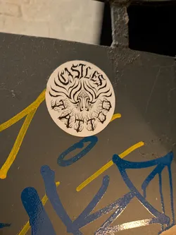 A circular white sticker featuring black artwork. The top curved text reads 'CASTLES' in a stylized gothic or blackletter font, and the bottom curved text reads 'TATTOO' in a spiky, flame-like font. In the center is an abstract, symmetrical organic tribal/flame motif resembling a spire or arrowhead.