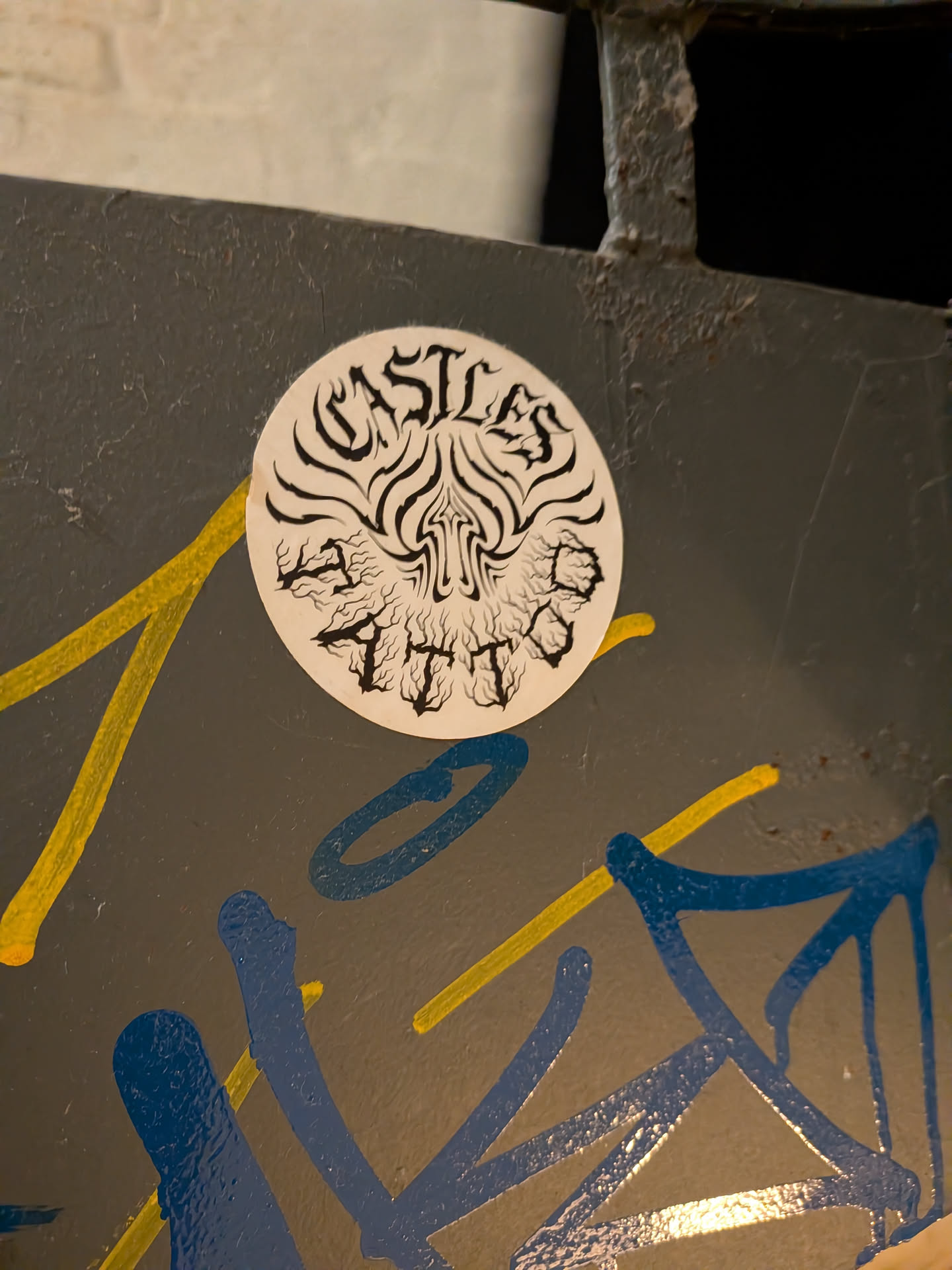 A circular white sticker featuring black artwork. The top curved text reads 'CASTLES' in a stylized gothic or blackletter font, and the bottom curved text reads 'TATTOO' in a spiky, flame-like font. In the center is an abstract, symmetrical organic tribal/flame motif resembling a spire or arrowhead.