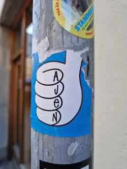 A blue and white sticker featuring a hand giving a thumbs up with the letters A, J, E, and N arranged vertically inside the hand's fingers. The sticker is affixed to a metal pole and shows some signs of wear and tear.