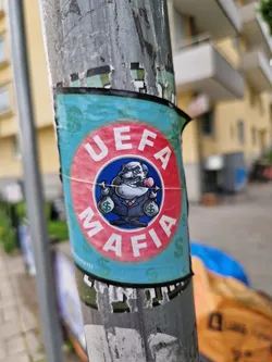 A sticker featuring a cartoon depiction of a man in a suit holding money bags, with the text "UEFA Mafia" written around him. The style is reminiscent of satirical political cartoons.