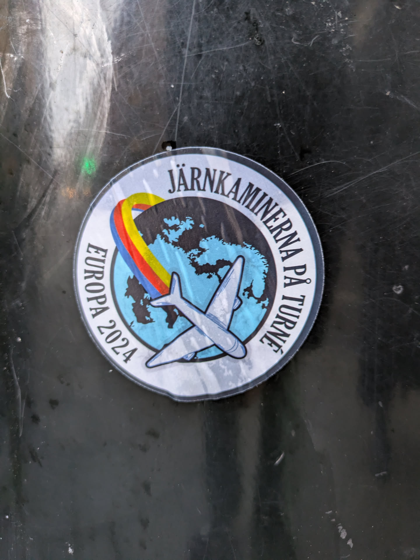 Round sticker featuring a plane flying over a world map with a rainbow arc. The text on the sticker says "Järnkaminerna på Europaturne 2024" which translates to "Iron Stoves on European Tour 2024".