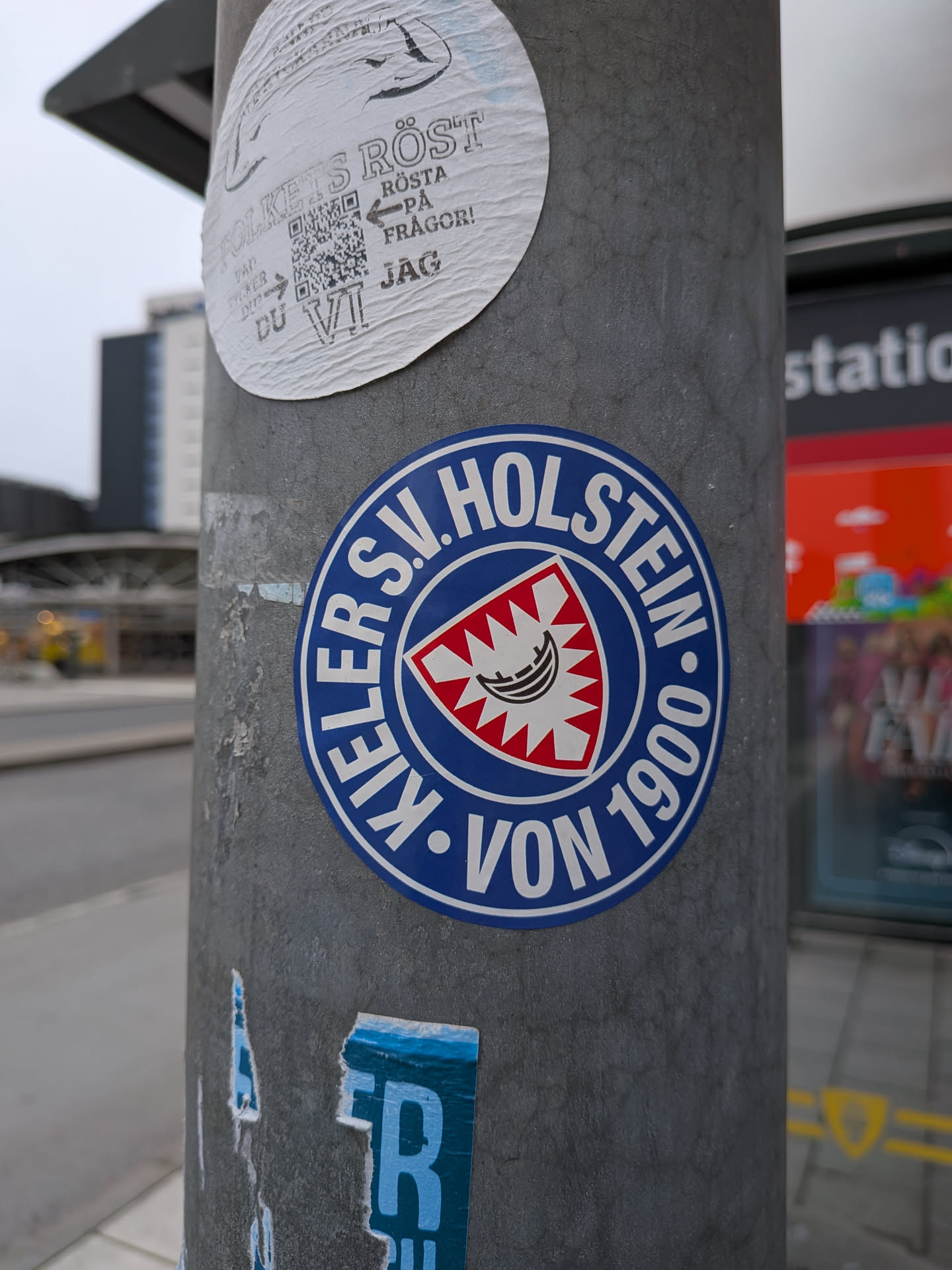 The sticker is circular with a prominent blue border. Inside the border, the text 'KIELER S.V. HOLSTEIN' is written in white capital letters, arranged in a semi-circle. Below this, 'VON 1900' is also in white capital letters. The center of the sticker features a shield with a red and white jagged pattern around the edges. Within this, a white emblem with black lines depicts a stylized boat or crescent shape, possibly representing a historical symbol of the region or club. The overall design is clean and emblematic, typical of sports club insignia.