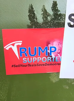 A red sticker with "TRUMP SUPPORTER" in blue text, using the Tesla logo as the "T" in "TRUMP". The sticker also features a portrait of Elon Musk and the hashtag "#SellYourTeslaSaveDemocracy".