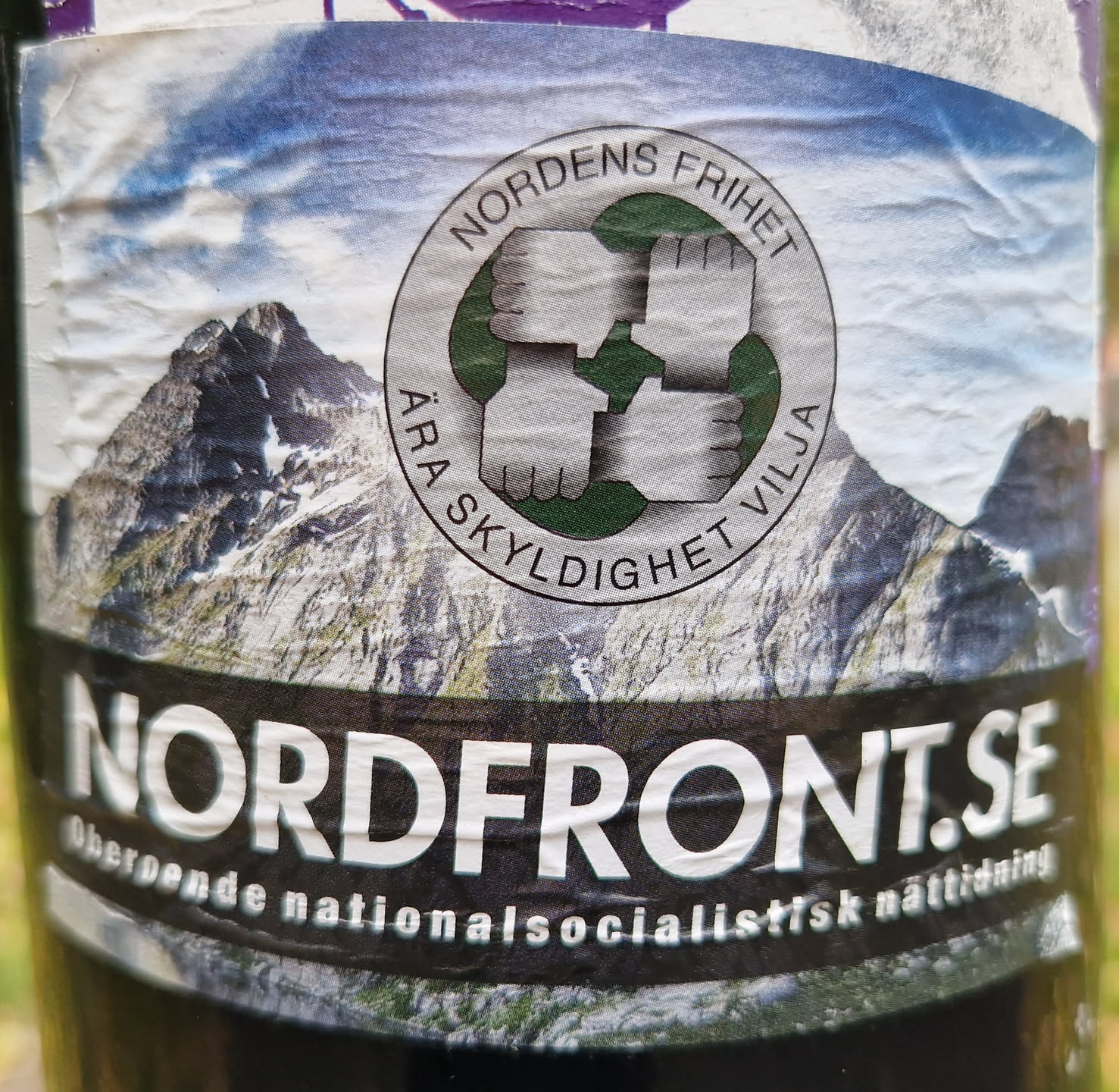 Sticker featuring a logo with four clenched fists in a circle, mountains in the background, and the text "Nordens Frihet " ("Freedom of the North") and "Ara Skyndighet Vilja" ("Race, Honor, Will") around the fists.  The bottom of the sticker displays the website address nordfront.se and describes it as an independent National Socialist online newspaper.