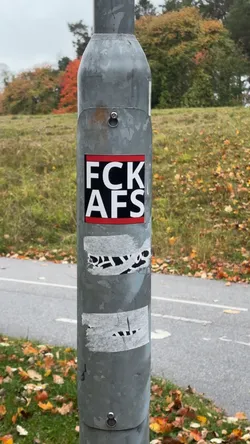 Street sticker A square-shaped sticker adhered to a galvanized metal pole. It features the bold white capitalized text 'FCK' on the top line and 'AFS' on the bottom line against a black central background. The design is framed by horizontal red bands at the very top and bottom, edged with a thin black border.