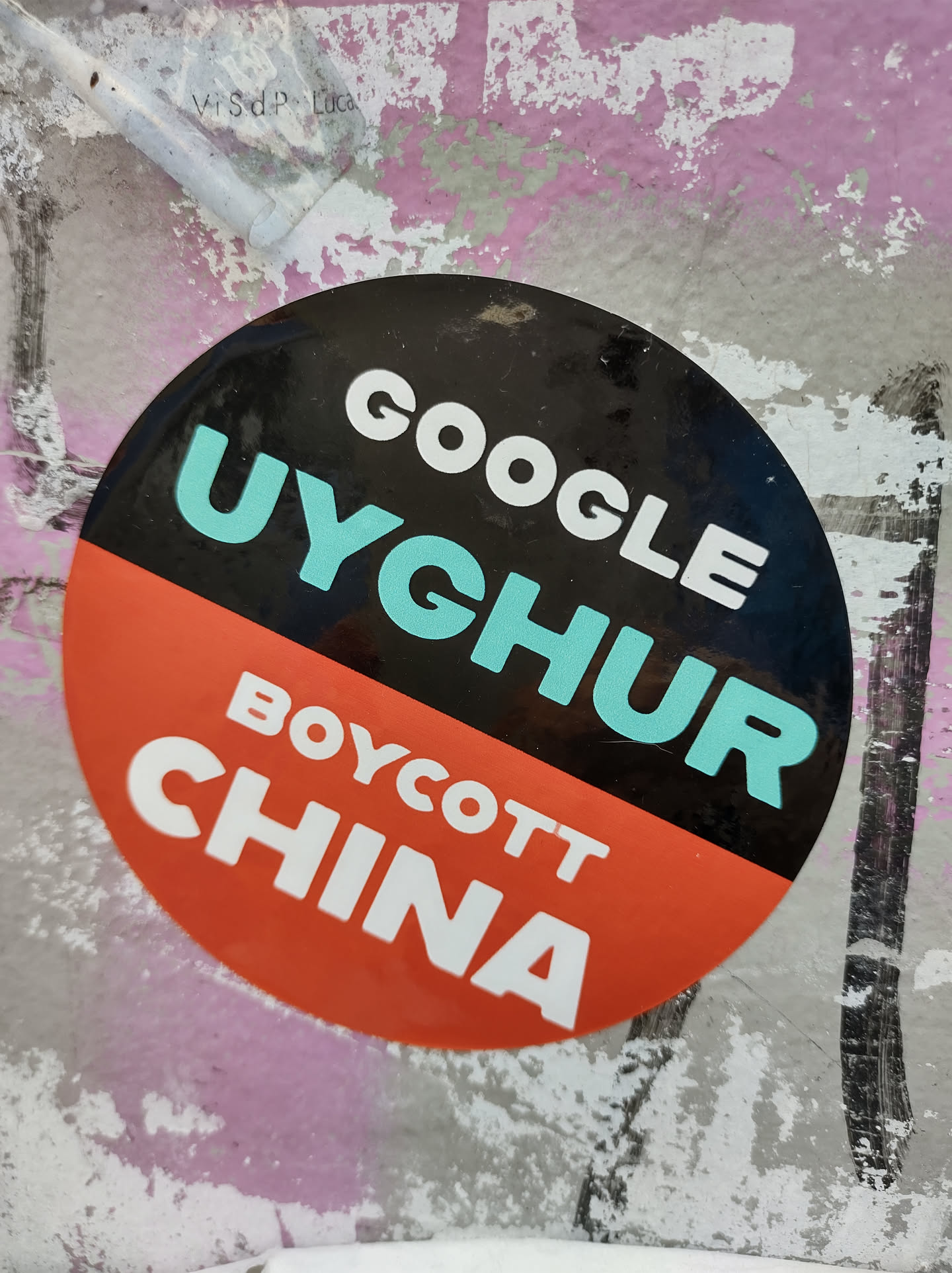 A round sticker with a black and orange split design. The top half is black with the words "GOOGLE UYGHUR" in white and teal. The bottom half is orange with the words "BOYCOTT CHINA" in white. The sticker appears to be affixed to a wall with some peeling paint.