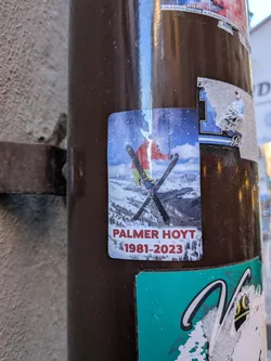 A vertical, rounded-corner photographic sticker featuring a skier captured mid-air executing a ski-crossing trick against a backdrop of snow-covered mountains and a partly cloudy sky. The skier wears a red/orange jacket, yellowish-green pants, and a white helmet. At the bottom of the image, printed over the snow in bold red capital letters, is the name 'PALMER HOYT' followed by the years '1981-2023'.